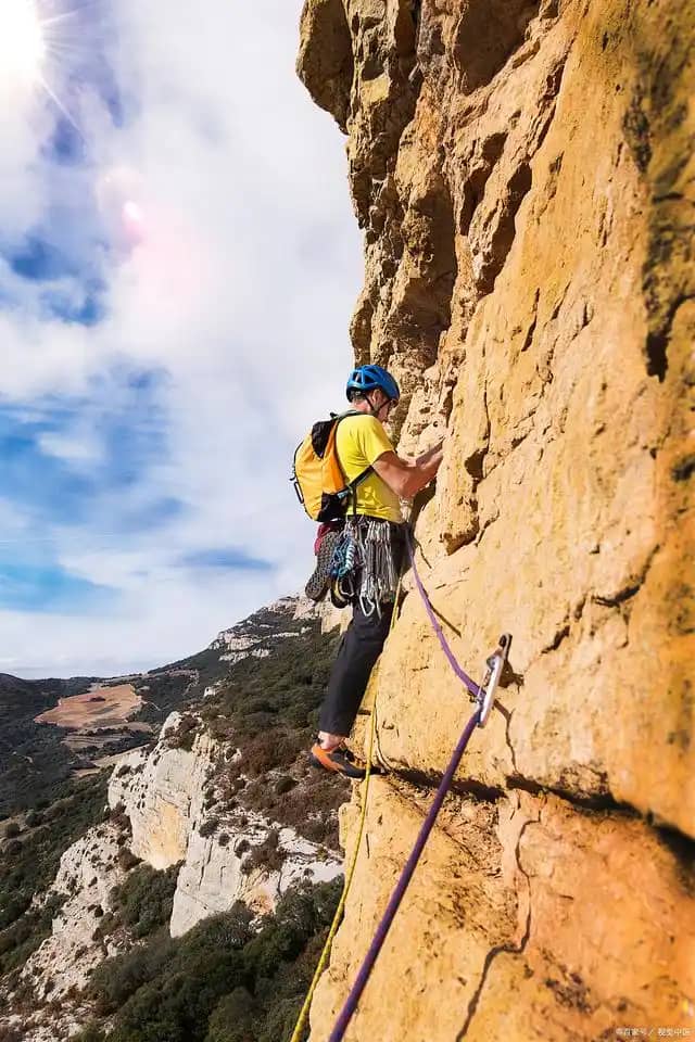 Rock climber scaling a steep cliff with safety gear against a scenic mountainous backdrop.