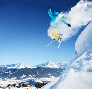 Skier performing a jump over snowy mountains under a clear blue sky, showcasing winter sports action.